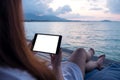 A woman holding and using black tablet with blank white desktop screen while sitting by the sea with blue sky background Royalty Free Stock Photo