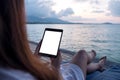 A woman holding and using black tablet with blank white desktop screen while sitting by the sea with blue sky background Royalty Free Stock Photo