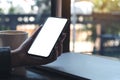 A woman`s hand holding black mobile phone with blank white desktop screen with coffee cup and laptop on table in cafe Royalty Free Stock Photo