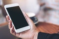 Mockup image of a woman`s hand holding white mobile phone with blank black desktop screen and laptop on wooden table Royalty Free Stock Photo