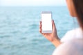 Mockup image of a woman`s hand holding white mobile phone with blank desktop screen by the sea and blue sky Royalty Free Stock Photo