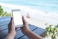 Mockup image of woman`s hand holding white mobile phone with blank desktop screen on wooden table in front of the sea and blue sky Royalty Free Stock Photo