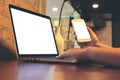 Woman`s hands holding mobile phone while using laptop with blank white screen on vintage wooden table in loft cafe Royalty Free Stock Photo