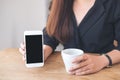 A woman`s hands holding and showing white mobile phone with blank black desktop screen with coffee cups on wooden table Royalty Free Stock Photo