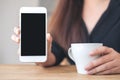 A woman`s hands holding and showing white mobile phone with blank black desktop screen with coffee cups on wooden table Royalty Free Stock Photo