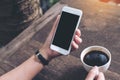 Mockup image of woman`s hands holding white mobile phone with blank black screen and white coffee cup on wooden table Royalty Free Stock Photo