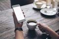 Mockup image of woman`s hands holding white mobile phone with blank screen and white coffee cup on wooden table Royalty Free Stock Photo