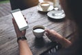 Mockup image of woman`s hands holding white mobile phone with blank screen and white coffee cup on wooden table Royalty Free Stock Photo
