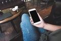 Woman`s hands holding white mobile phone with blank screen on thigh and laptop and coffee cup on wooden table in cafe Royalty Free Stock Photo