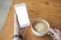 Mockup image of woman`s hands holding white mobile phone with blank screen and white coffee cup of hot latte on wooden table Royalty Free Stock Photo