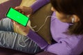 Mockup image of woman on sofa and holding mobile phone with headphones and blank green screen