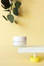Mockup moisturizer cream in jar standing on abstract pedestal on pastel yellow background with copy space and defocused eucalyptus