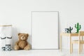 Mockup poster, teddy bear and material basket placed on the floor in white room interior with wooden table and small chair. Paste