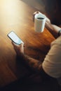 Mockup shot of man`s hand with tattoos holding  cell phone and coffee cup with blank screen on desk at home office Royalty Free Stock Photo