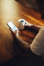 Mockup shot of man`s hand with tattoos holding  cell phone and coffee cup with blank screen on desk at home office Royalty Free Stock Photo