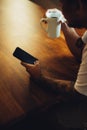 Mockup shot of man`s hand with tattoos holding  cell phone and coffee cup with blank screen on desk at home office Royalty Free Stock Photo