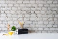 Mockup table with cup of coffee, book, wooden dummy model and jar of pencil on white table and brick wall background.