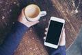 Mockup top view image of woman`s hands holding white mobile phone with blank black screen and a coffee cup on wooden table Royalty Free Stock Photo