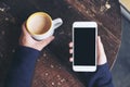 Mockup top view image of woman`s hands holding white mobile phone with blank black screen and a coffee cup on wooden table Royalty Free Stock Photo