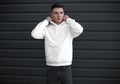 Mockup white hoodie with a pocket on a guy, straightening a hood, front view, men`s blank long sleeve clothes against a metal wal