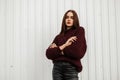 Model of a beautiful young woman in a stylish burgundy knitted sweater in leather trendy black pants posing on the street near a