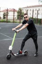 Model of a cool young man in a black clothes in sunglasses in sneakers stands with an electric scooter on an open basketball court Royalty Free Stock Photo