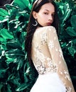 Model with dark long hair in wide-leg classic pants posing near green tropical exotic leaves