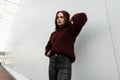 Model of a fashionable young woman in a stylish burgundy sweater in leather trendy black pants posing on the street near a white