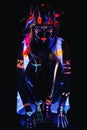 Model in image of Native american with neon makeup, which made of fluorescent paint in ultraviolet light.