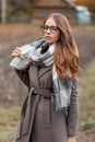 Model of a stylish young hipster woman in a fashionable coat in trendy glasses in a knitted vintage scarf walks in the countryside Royalty Free Stock Photo