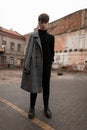Model of a stylish young man in an autumn fashionable gray check coat in black trendy clothing in vintage leather shoes stands on