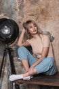 Model of a stylish young woman with blond hair in a beige t-shirt in blue jeans enjoys relaxation near a vintage brick wall Royalty Free Stock Photo