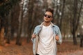 Model trendy American young man in fashionable sunglasses in a stylish denim jacket with a hairstyle in the park. Handsome guy Royalty Free Stock Photo