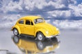 Model of a yellow Beetle parked on a puddle and the lights on, we can see its reflection on the puddle and a nice cloudy sky