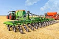 Agricultural seeders attached to tractors stand on yellow stubble in a wide field against a blue cloudy sky on a summer day