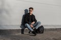 Modern American young hipster man in fashionable sunglasses in stylish casual denim clothes in sneakers with backpack is rests Royalty Free Stock Photo