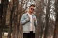 Modern American young hipster man in a stylish denim jacket in a white vintage t-shirt in trendy sunglasses posing in the autumn Royalty Free Stock Photo