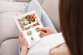 Modern app for ordering meal at home. Woman in casual sitting on sofa and using tablet to choose takeaway food