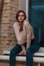 Modern attractive girl with brown long hair in a pink sweatshirt in vintage jeans sits on a windowsill near a brick wall.