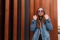 Modern attractive young woman in stylish blue denim jacket in military hoodie with fashionable purple glasses posing near a metal Royalty Free Stock Photo