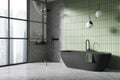 Modern bathroom with black bathtub and shower, green tiled wall, and large windows. Minimalist decor. Light, cityscape background