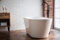 Modern bathroom with freestanding white acrylic bathtub and black loft mixer. Walls made of bricks, parquet is wooden on floor,
