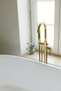 Modern bathroom with stylish golden faucet, white bathtub and travertine tiles. Stylish bathroom details with summer flowers