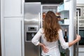 Modern beautiful long haired woman standing and open refrigerators door indoors and looking forward inside the fridge.