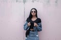 Modern beautiful young hipster woman in stylish black sunglasses stands and straightens a denim fashion jacket near a vintage wall Royalty Free Stock Photo