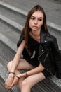 Modern beautiful young woman in a vintage leather jacket in a stylish black dress sits on the steps in the city. Elegant modern