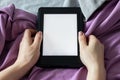 A modern black e-reader electronic book with a blank screen in female hands on a gray and purple bed. Mockup tablet closeup