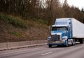 Modern blue powerful stylish big rig semi truck with semi trailer going on wide highway with trees on the hillside shoulder Royalty Free Stock Photo