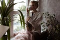 Modern, calm, pensive blond woman in glasses and warm clothes sitting on windowsill and drink coffee or tea with blanket Royalty Free Stock Photo