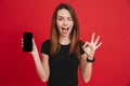 Modern caucasian woman rejoicing and showing ok sign while advertising new cell phone, isolated over red background